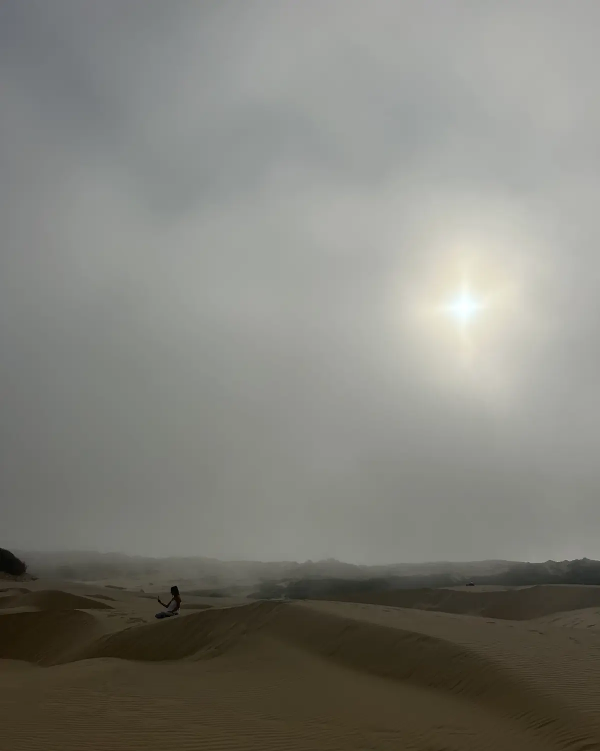 A small child sitting on a sand dune, reaching up toward a bright sun shining through the mist.